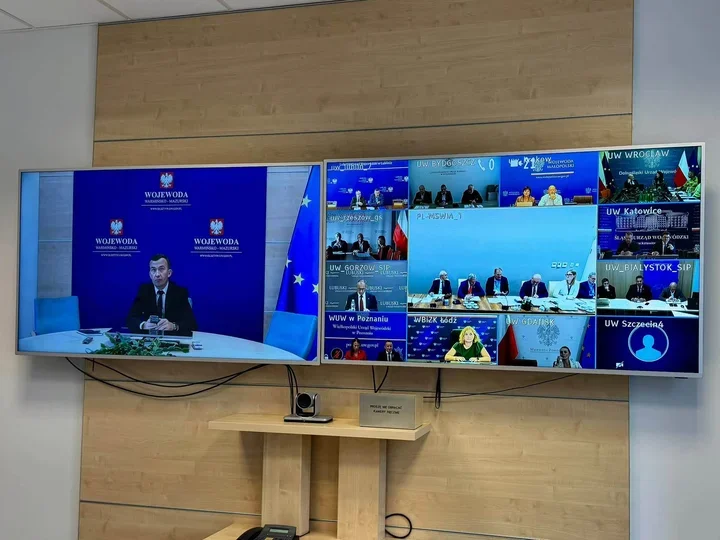Wideokonferencja z udziałem wojewodów i urzędów wojewódzkich, wyświetlana na dwóch dużych ekranach w sali konferencyjnej.