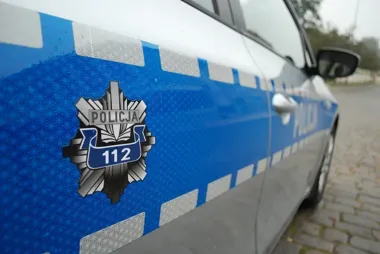 Fragment oznakowanego policyjnego radiowozu z emblematem Policji i numerem 112 zaparkowanego na brukowanej nawierzchni.