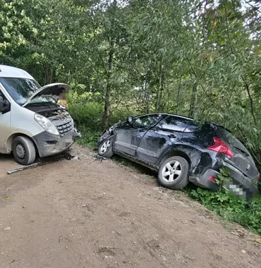 Dwa rozbite samochody, biały van i czarny osobowy, stoją na leśnej drodze po zderzeniu.