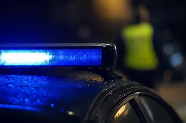 Nocny zbliżenie na niebieski sygnał świetlny na dachu policyjnego radiowozu, w tle rozmyta postać w odblaskowej kamizelce.