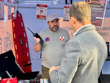 Mężczyzna w szarej koszulce z emblematem ratunkowym pokazuje przenośne radio innemu mężczyźnie podczas plenerowej prezentacji sprzętu ratowniczego.
