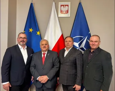 Czterech mężczyzn w garniturach pozuje do zdjęcia przed flagami Unii Europejskiej, Polski i NATO oraz godłem państwowym.