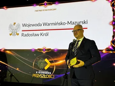 Mężczyzna w garniturze stoi przy mikrofonie na scenie podczas X Jubileuszowego Kongresu Przyszłości, trzymając nagrodę, na tle ekranu z napisem „Wojewoda Warmiń