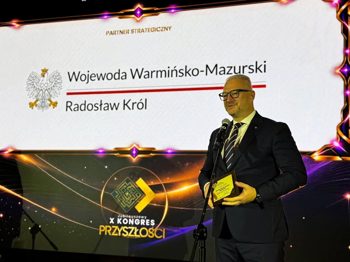 Mężczyzna w garniturze stoi przy mikrofonie na scenie podczas X Jubileuszowego Kongresu Przyszłości, trzymając nagrodę, na tle ekranu z napisem „Wojewoda Warmiń