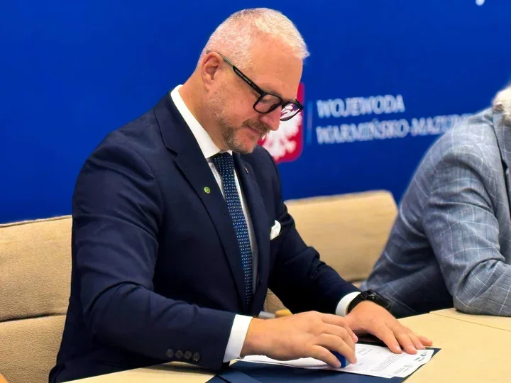 Mężczyzna w garniturze siedzi przy stole i podpisuje dokument, w tle niebieska ścianka z napisem „Wojewoda Warmińsko-Mazurski”.
